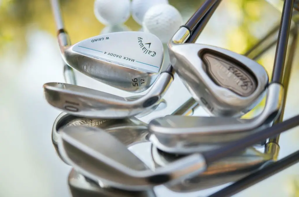 A close-up of a selection of golf clubs and a golf ball in a bag, with the focus on the shiny metal surfaces of the club heads. The details and grooves on the irons are clearly visible, highlighting the brands and loft angles, such as '56 degree' and '10 iron'.